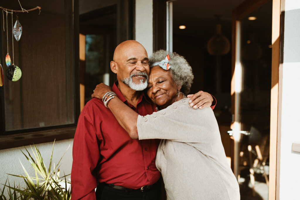 Smiling elderly woman embracing husband at front yard. Smiling senior couple is standing with eyes closed. They are in casuals.