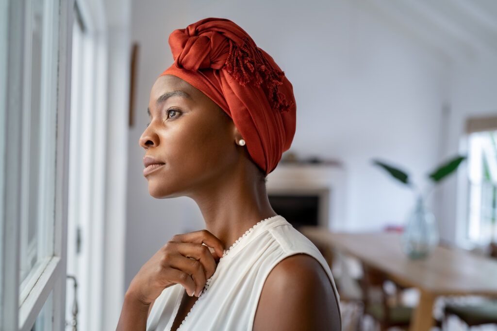 Beautiful pensive woman wearing headscarf looking outside window in contemplation. Mature black woman wearing a traditional turban and standing near the window at home while thinking. Worried African mature woman with cancer at home.