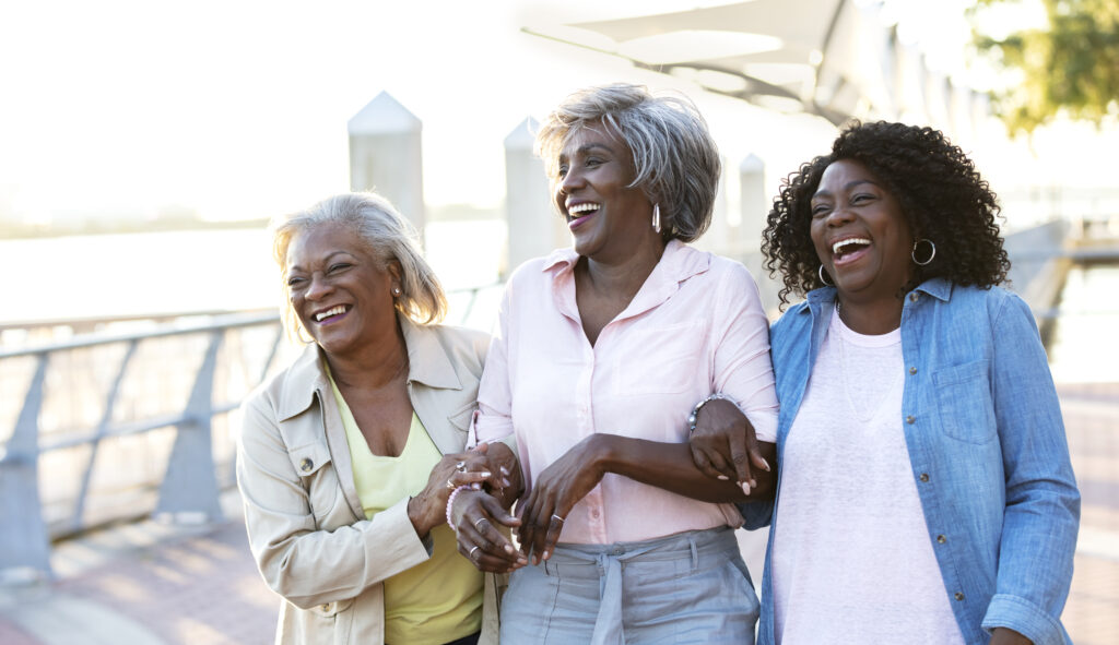 A group of three senior African-American women, best friends, hanging out together on a sunny day on a city waterfront. They are walking arm in arm, conversing and laughing, having a good time. The woman in the middle is in her 70s and her friends are in their 60s.